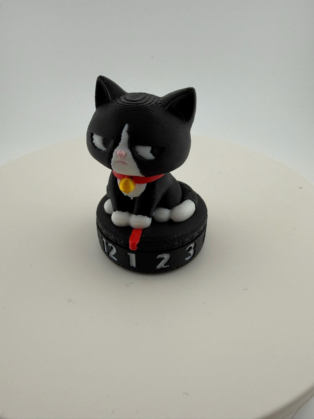 May include: A black and white cat figurine with a red collar and a gold bell. The cat sits atop a black circular base with the numbers 1, 2, 3, and 12. The figurine is a decorative item.