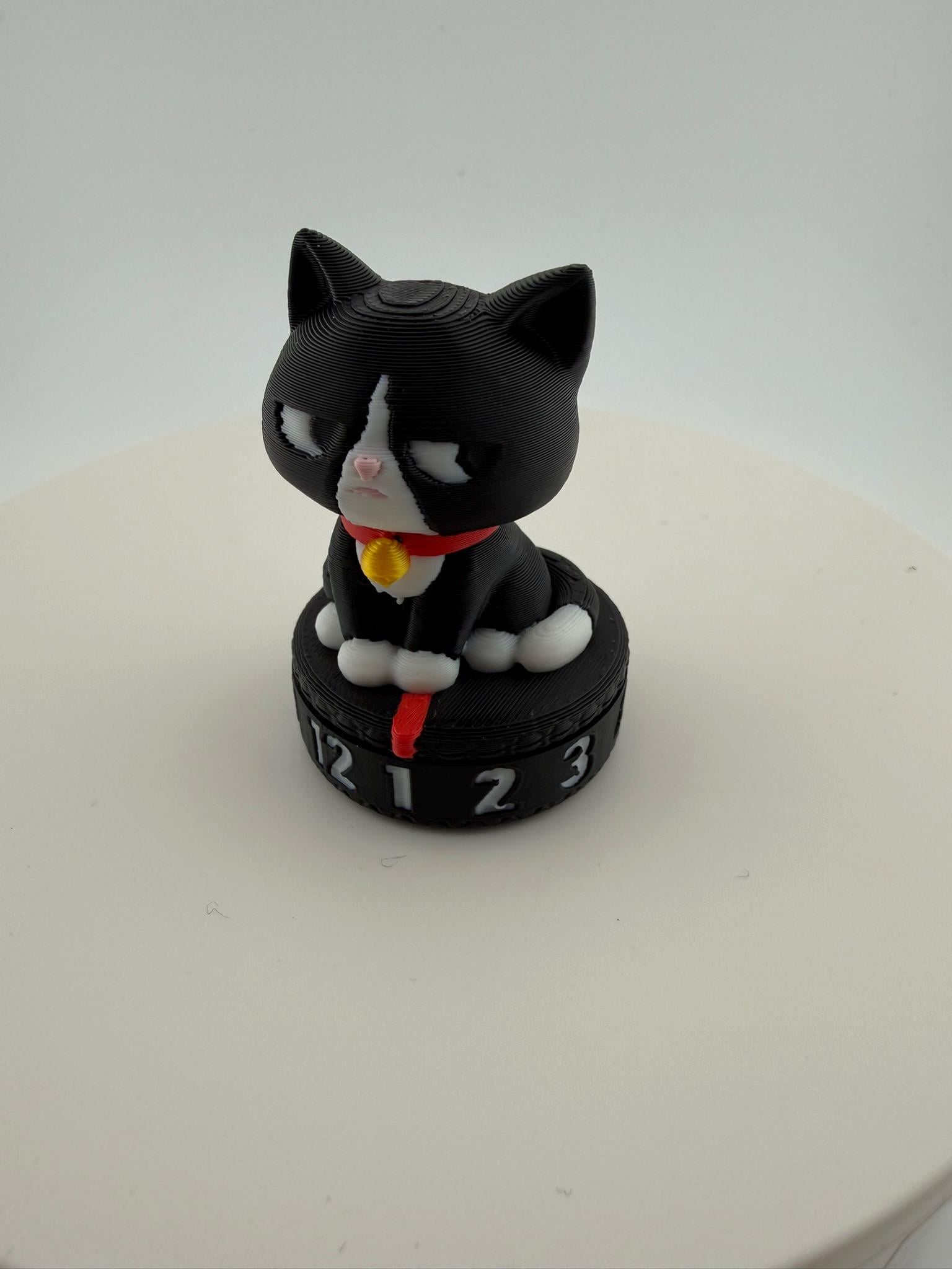 May include: A black and white cat figurine with a red collar and a gold bell. The cat sits atop a black circular base with the numbers 1, 2, 3, and 12. The figurine is a decorative item.