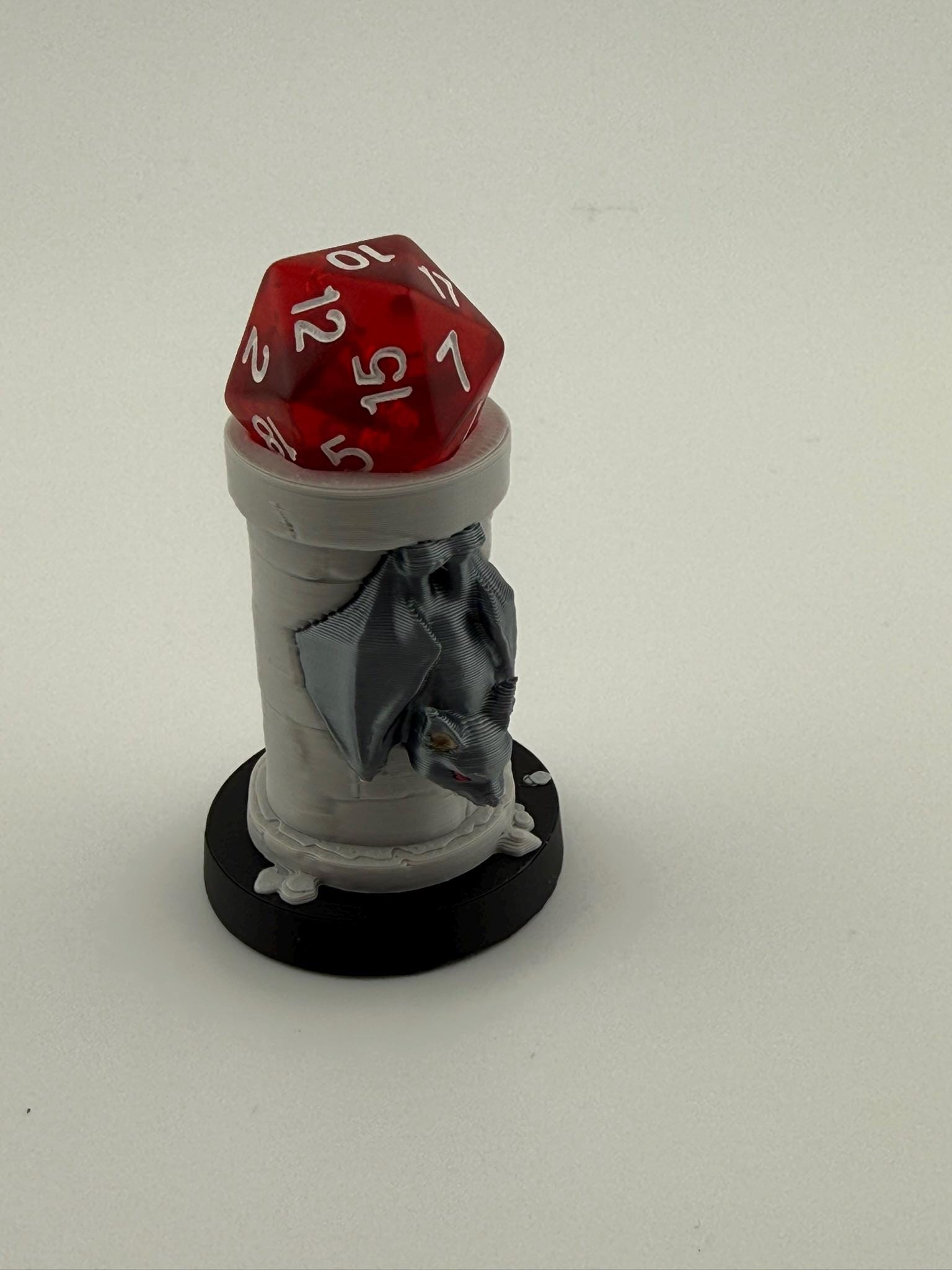May include: A red, multi-sided die rests atop a white, cylindrical dice tower with a gray bat-like figure attached. The tower sits on a black circular base. The die has white numbers.