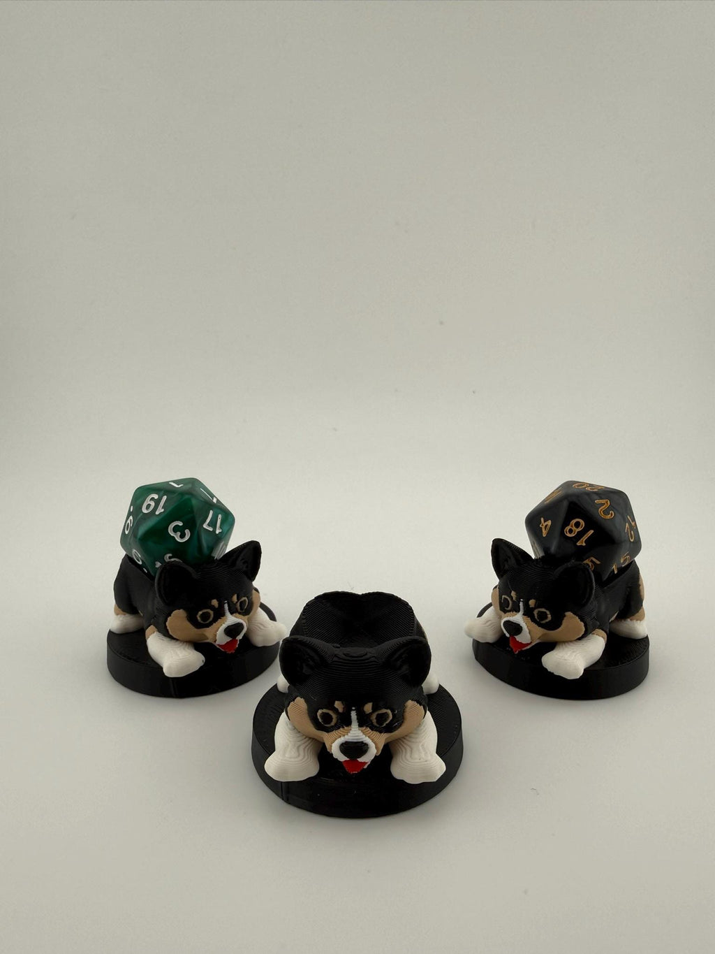 May include: Three black and white corgi-shaped dice holders with red tongues, each on a black circular base. Two holders have a multi-sided die on top. The dice are green and black with gold numbers. The holders are for tabletop games.