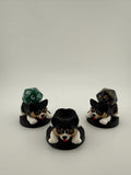 May include: Three black and white corgi-shaped dice holders with red tongues, each on a black circular base. Two holders have a multi-sided die on top. The dice are green and black with gold numbers. The holders are for tabletop games.