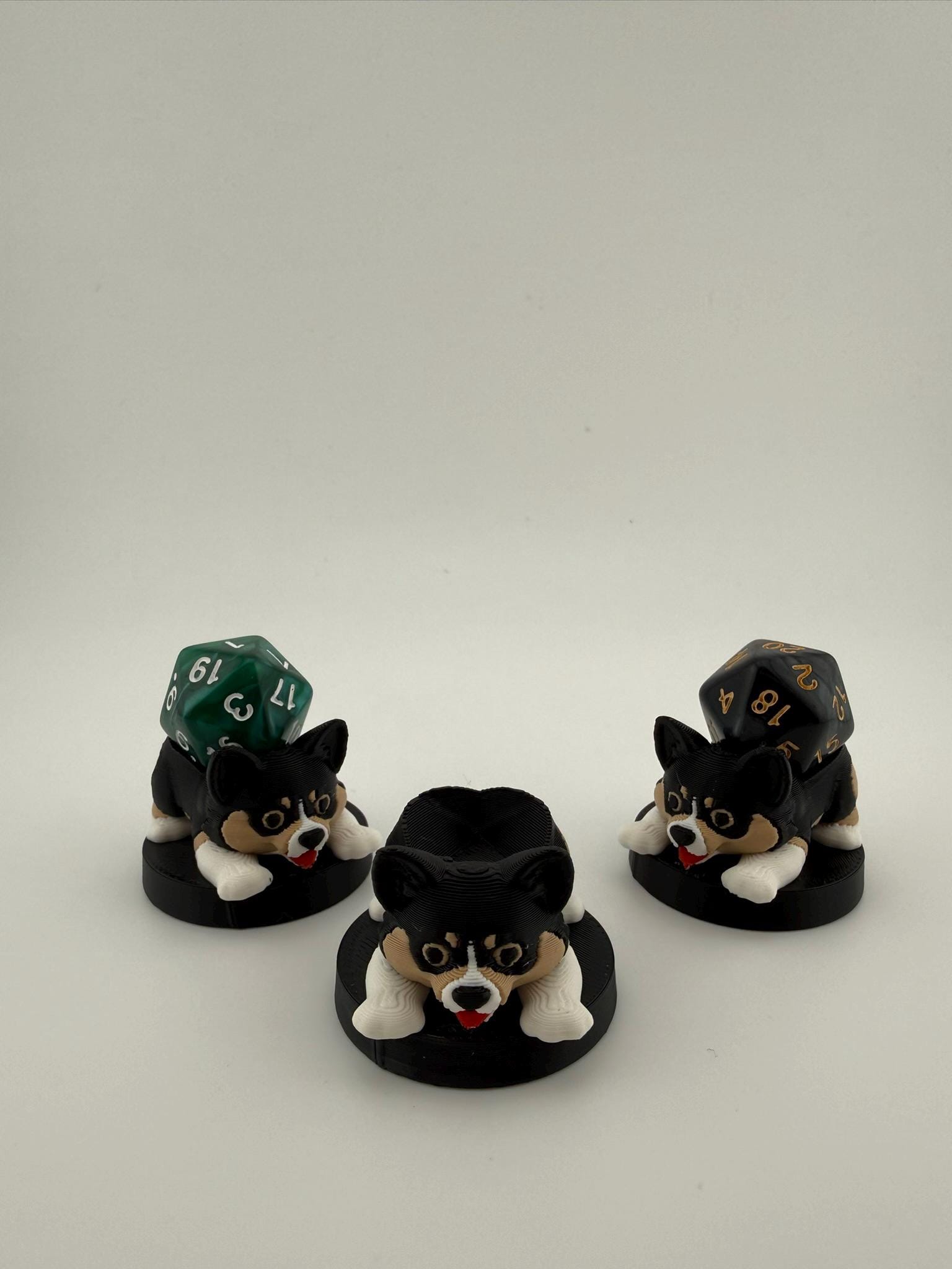 May include: Three black and white corgi-shaped dice holders with red tongues, each on a black circular base. Two holders have a multi-sided die on top. The dice are green and black with gold numbers. The holders are for tabletop games.