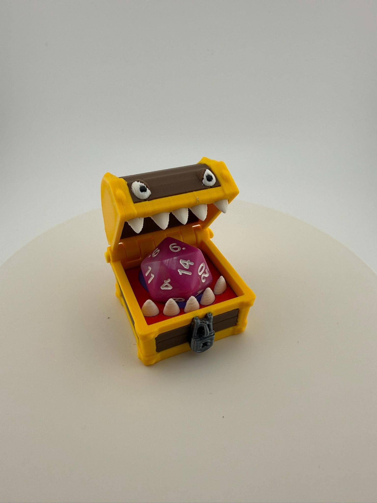 May include: A yellow treasure chest with a brown lid and white teeth, holding a large, purple, twenty-sided die. The chest has a small, gray lock and is set against a white background. A fun item for tabletop gaming.