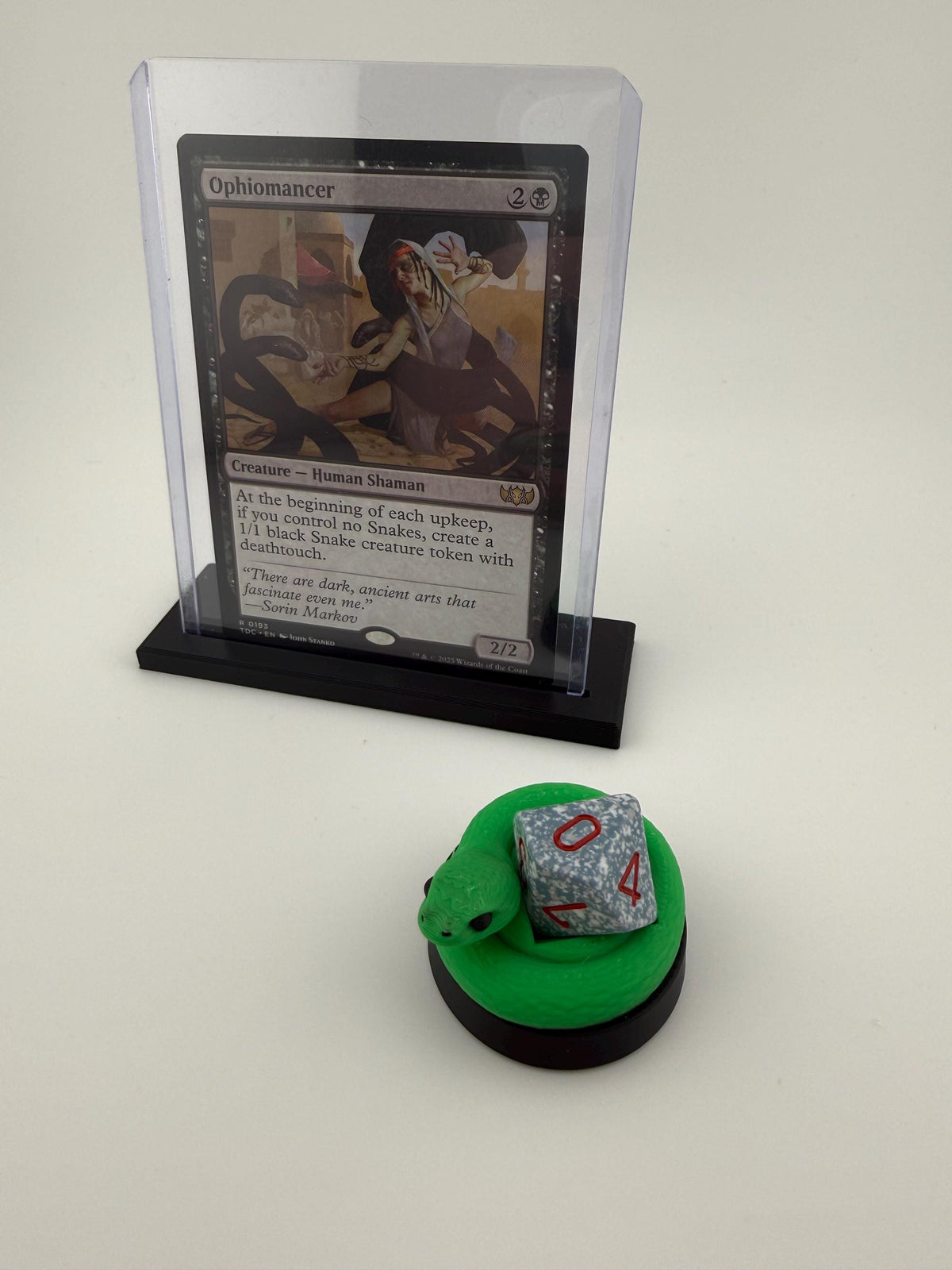 May include: A Magic: The Gathering card titled "Ophiomancer" in a clear plastic holder, displayed on a black stand. A green snake-shaped dice holder with a gray die showing the number 4 is in front of the card. The card features a black and white illustration.