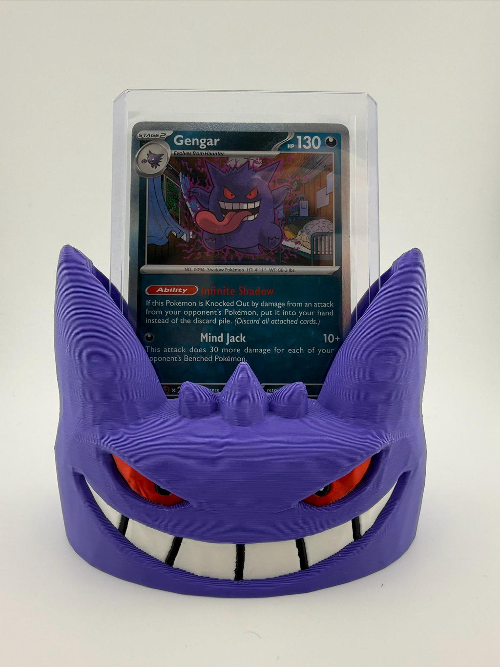 May include: A purple 3D-printed display stand shaped like the face of the character Gengar, featuring red eyes and a wide white grin. The stand holds a clear plastic protective case containing a Gengar trading card with 130 HP and text.