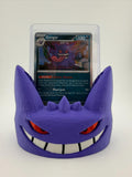 May include: A purple 3D-printed display stand shaped like the face of the character Gengar, featuring red eyes and a wide white grin. The stand holds a clear plastic protective case containing a Gengar trading card with 130 HP and text.